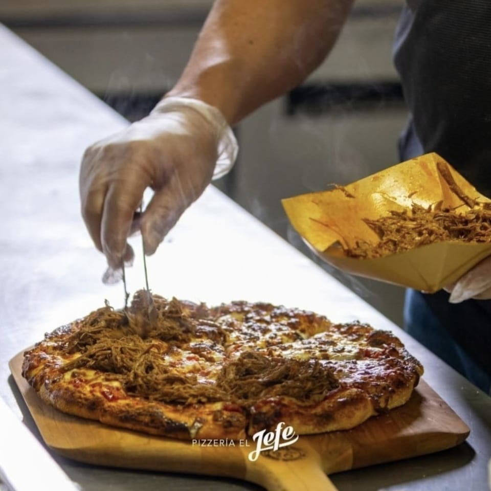 Gloved chef adds shredded meat to a cheese-covered pizza on a wooden board at Pizzería El Jefe.
