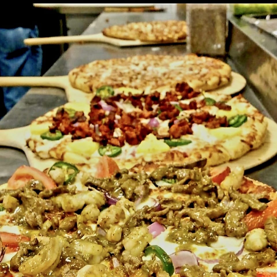 Several pizzas on wooden peels in a commercial kitchen, topped with cheese, seasoned meats, peppers, onions, and tomatoes.