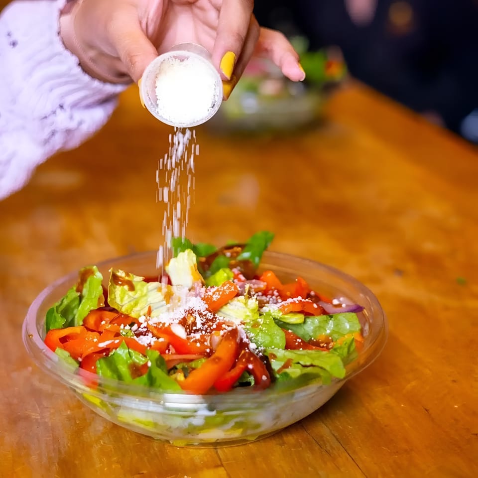 Fresh garden salad with lettuce, tomatoes, cucumbers, avocado, and feta cheese being sprinkled with seasoning.