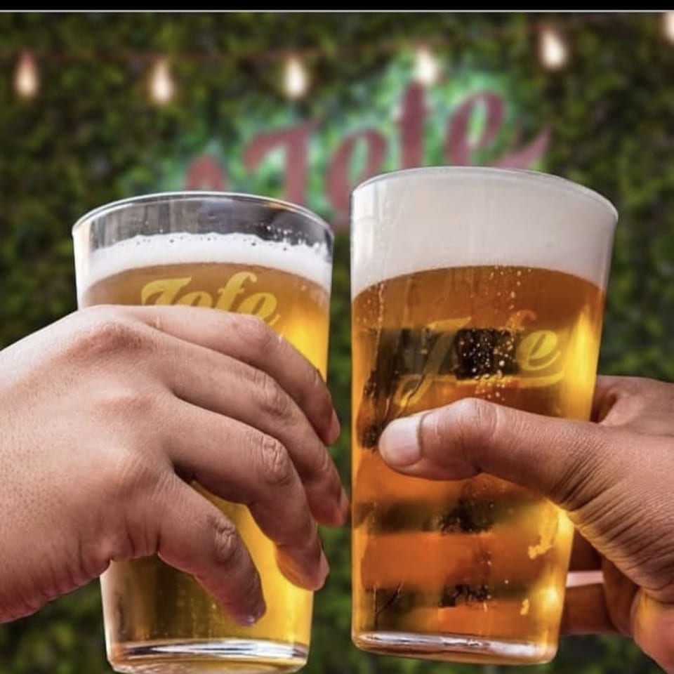 Two hands raise branded pint glasses of golden beer with foamy heads against a leafy outdoor background.