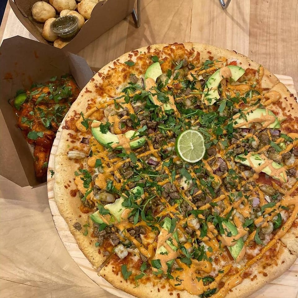 Loaded pizza topped with beef, mushrooms, avocado, cilantro, lime, and creamy sauce, alongside saucy wings and potatoes.