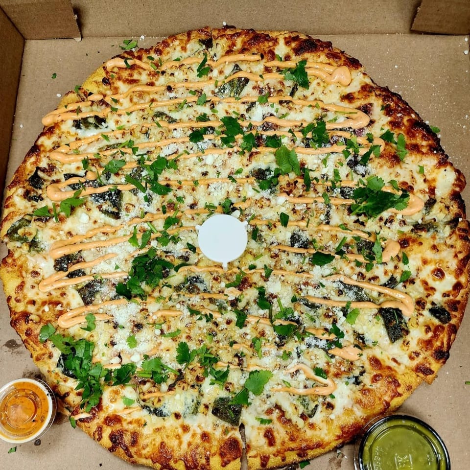 Large loaded pizza in a cardboard box, topped with melted cheese, herbs, creamy orange sauce, and dipping sauces.