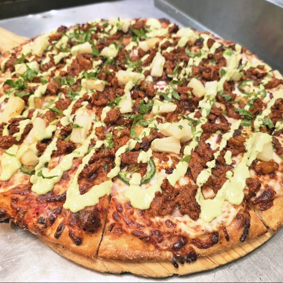 Round pizza topped with seasoned meat, pineapple chunks, jalapeños, cilantro, and drizzled green sauce on a wooden serving board.