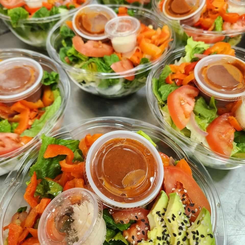 Multiple clear containers hold fresh salads with lettuce, tomatoes, peppers, avocado, sesame seeds, and individual dressing cups.
