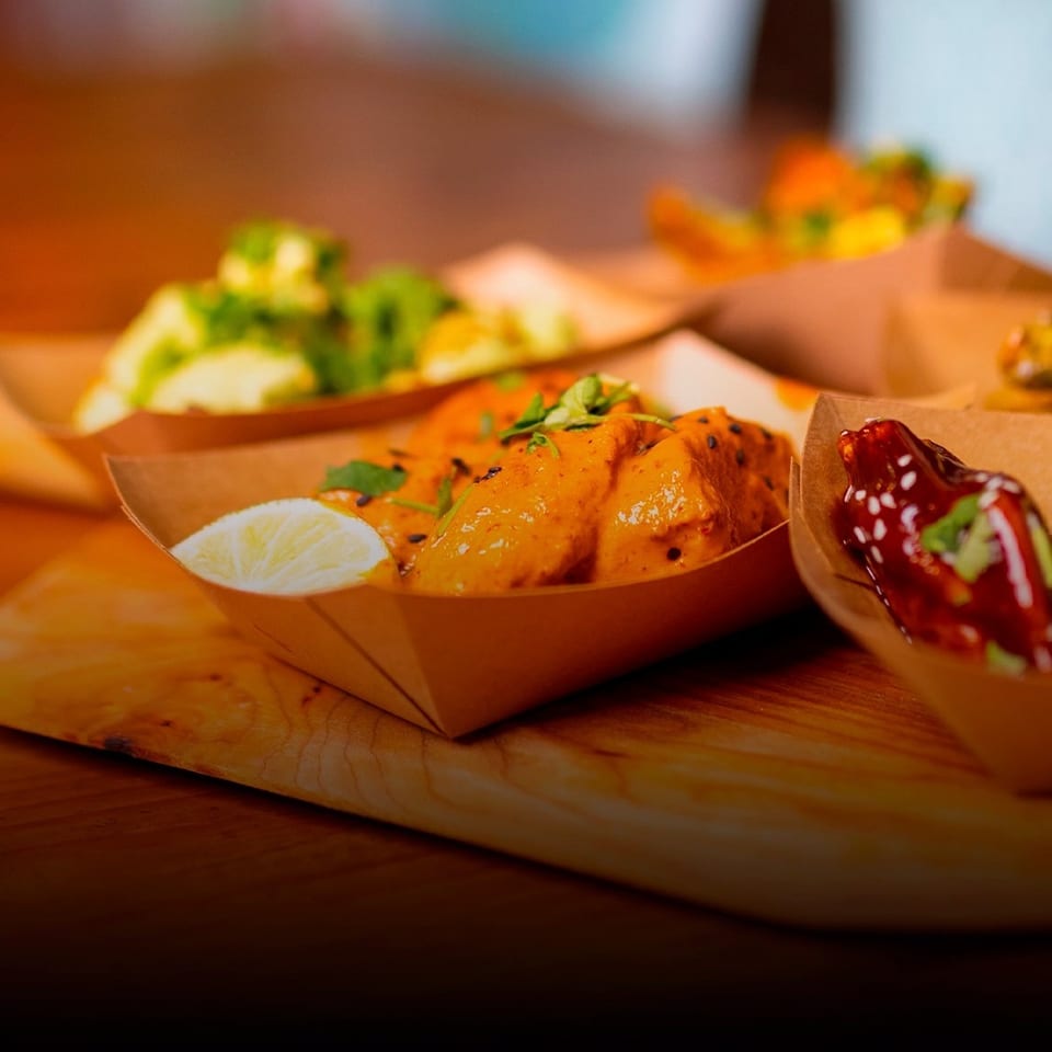Street-food trays hold glazed orange chicken with lime, salad, and a glossy red-sauced dish on a wooden table.