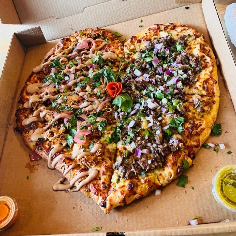 Two loaded pizzas in a cardboard box topped with cheese, chopped meat, herbs, onions, pickled vegetables, and drizzled sauce.