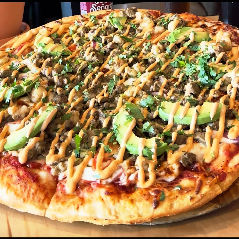 Large pizza topped with mushrooms, avocado, chopped herbs, melted cheese, and drizzled orange sauce on a wooden table.
