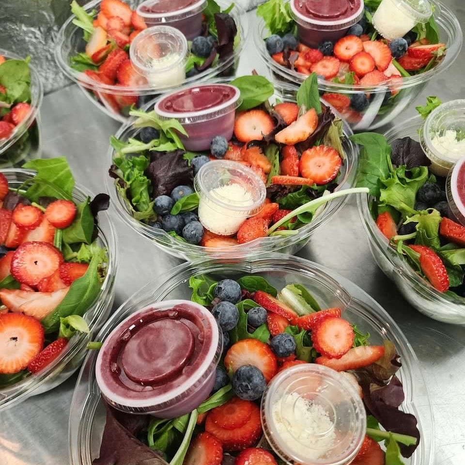 Clear containers of mixed green salads topped with strawberries and blueberries, accompanied by small cups of creamy dressing and berry sauce.