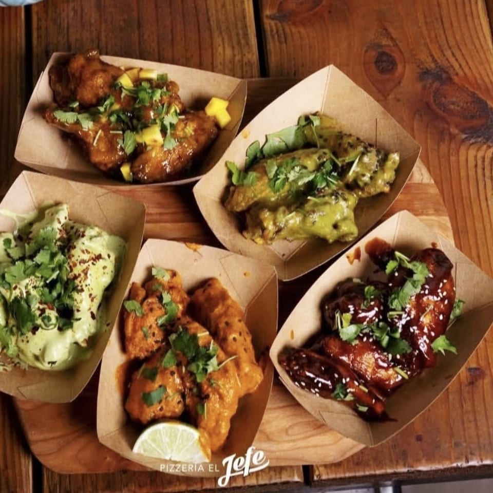 Assorted saucy chicken wings and tenders garnished with cilantro and sesame seeds in paper trays on a wooden table.
