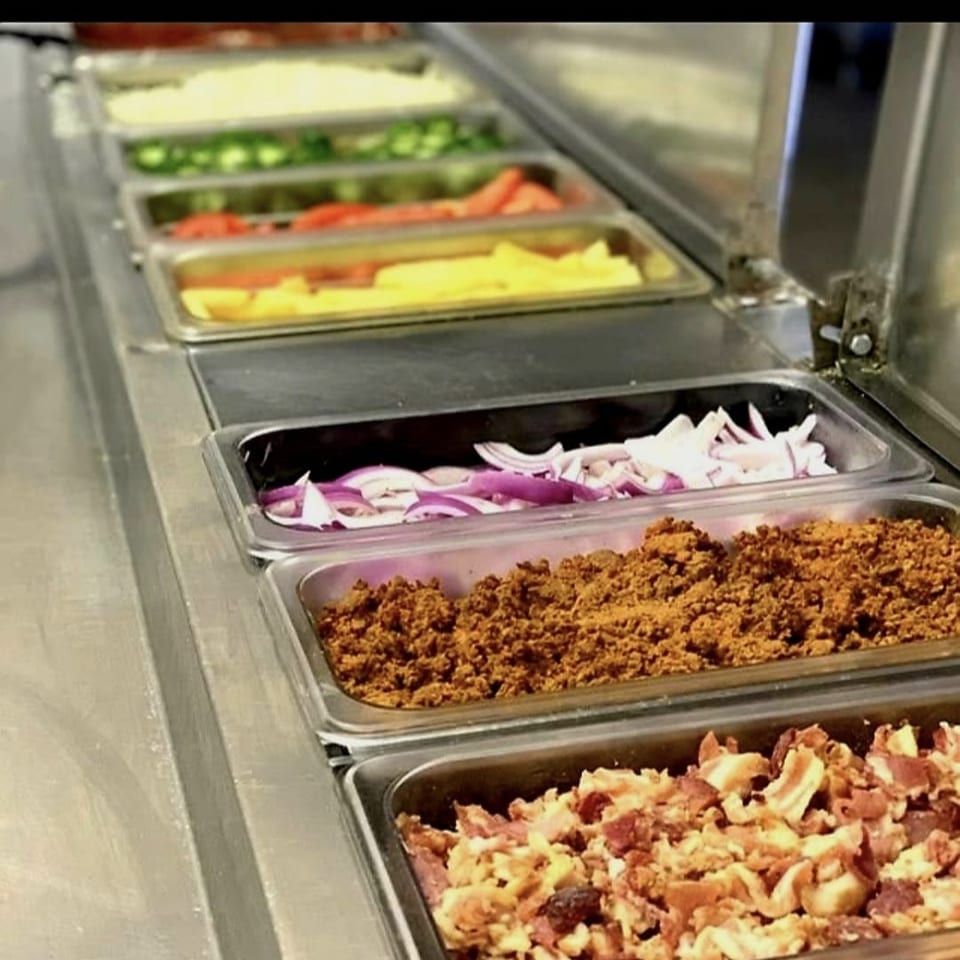 Stainless-steel buffet line displaying sliced vegetables, red onions, seasoned ground meat, and chopped bacon toppings.