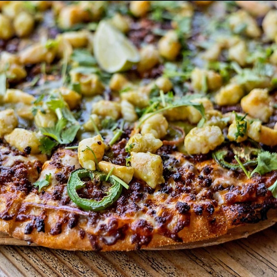 Close-up of a crispy flatbread topped with melted cheese, browned cauliflower, herbs, lime, and sliced jalapeño.